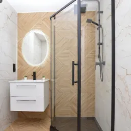 Nowoczesna łazienka z kabiną prysznicową typu walk-in, umywalką nablatową z szafką w kolorze białym, okrągłym lustrem z podświetleniem LED i grzejnikiem drabinkowym w kolorze czarnym, wykończona...