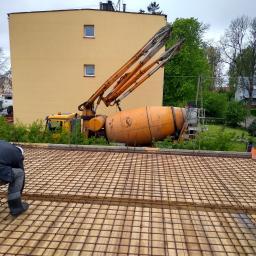 WSK Service - Pracownik w czarnej kurtce i gumowcach układa siatkę zbrojeniową na deskowaniu, w tle pompa do betonu i żółty budynek mieszkalny.