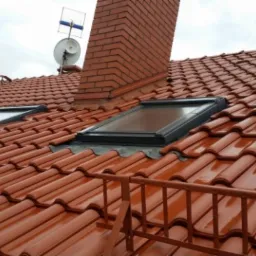 Czerwona dachówka ceramiczna, dwa okna dachowe z czarną ramą, ceglany komin z anteną satelitarną i telewizyjną na szczycie, widoczne z poziomu dachu z fragmentem barierki.