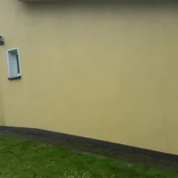 Żółta elewacja domu z widocznymi zaciekami i zabrudzeniami, oparta o ścianę drabina, trawnik w dolnej części kadru.