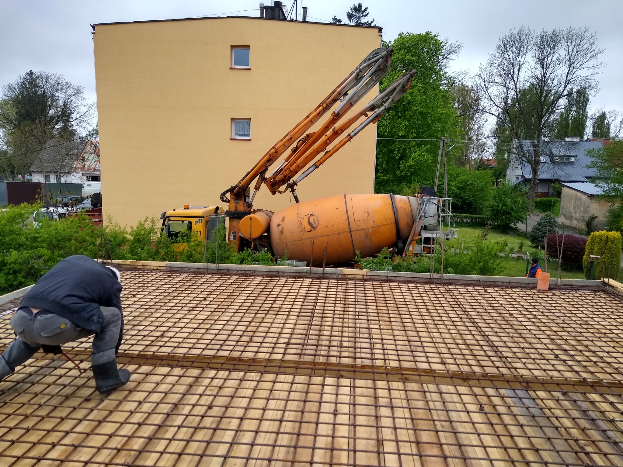 Pracownik w czarnej kurtce i gumowcach układa siatkę zbrojeniową na deskowaniu, w tle pompa do betonu i żółty budynek mieszkalny.