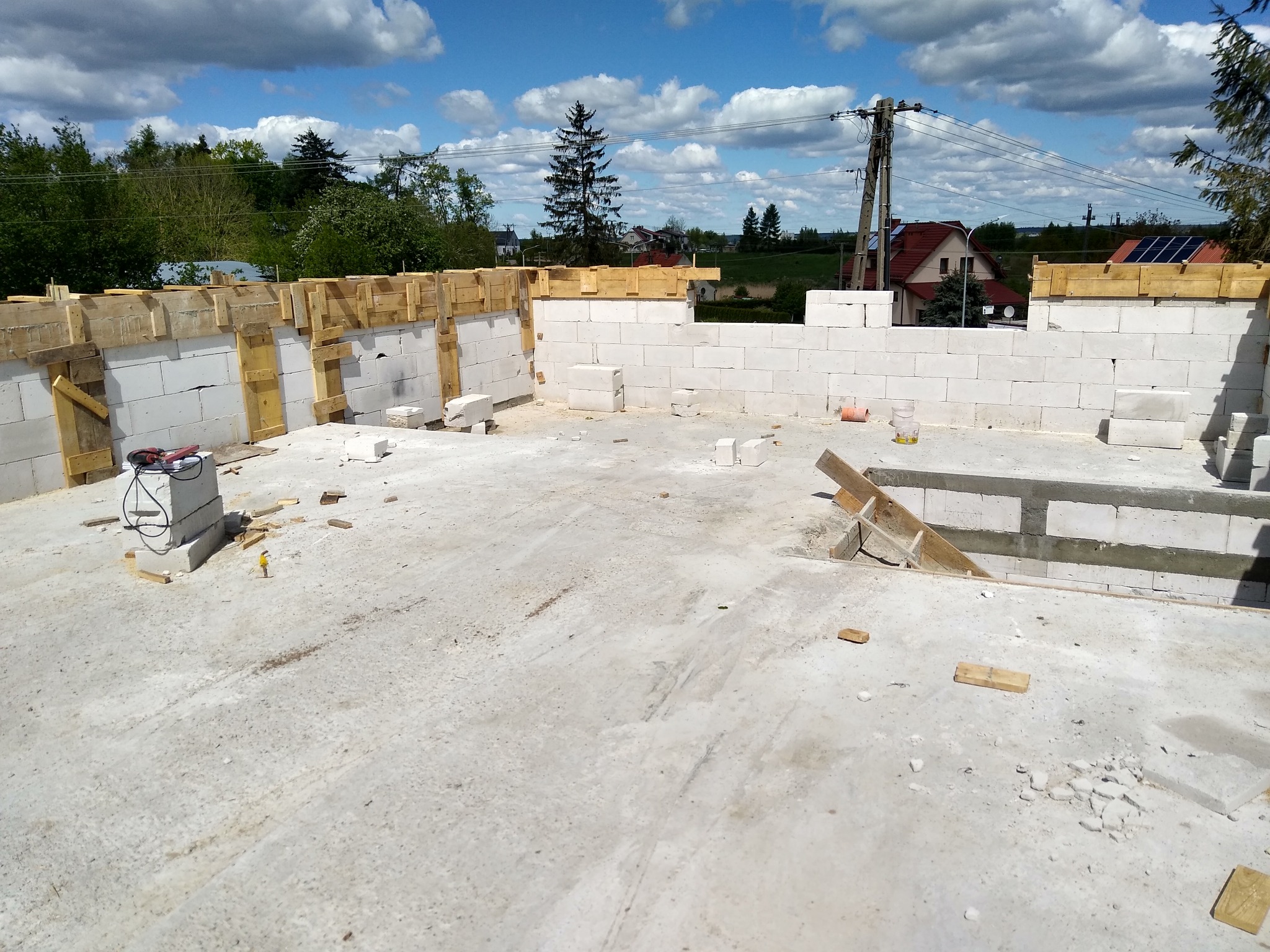 Wznoszone ściany z bloczków betonowych z drewnianym szalunkiem, widoczne narzędzia budowlane i fragment krajobrazu z domami w tle.