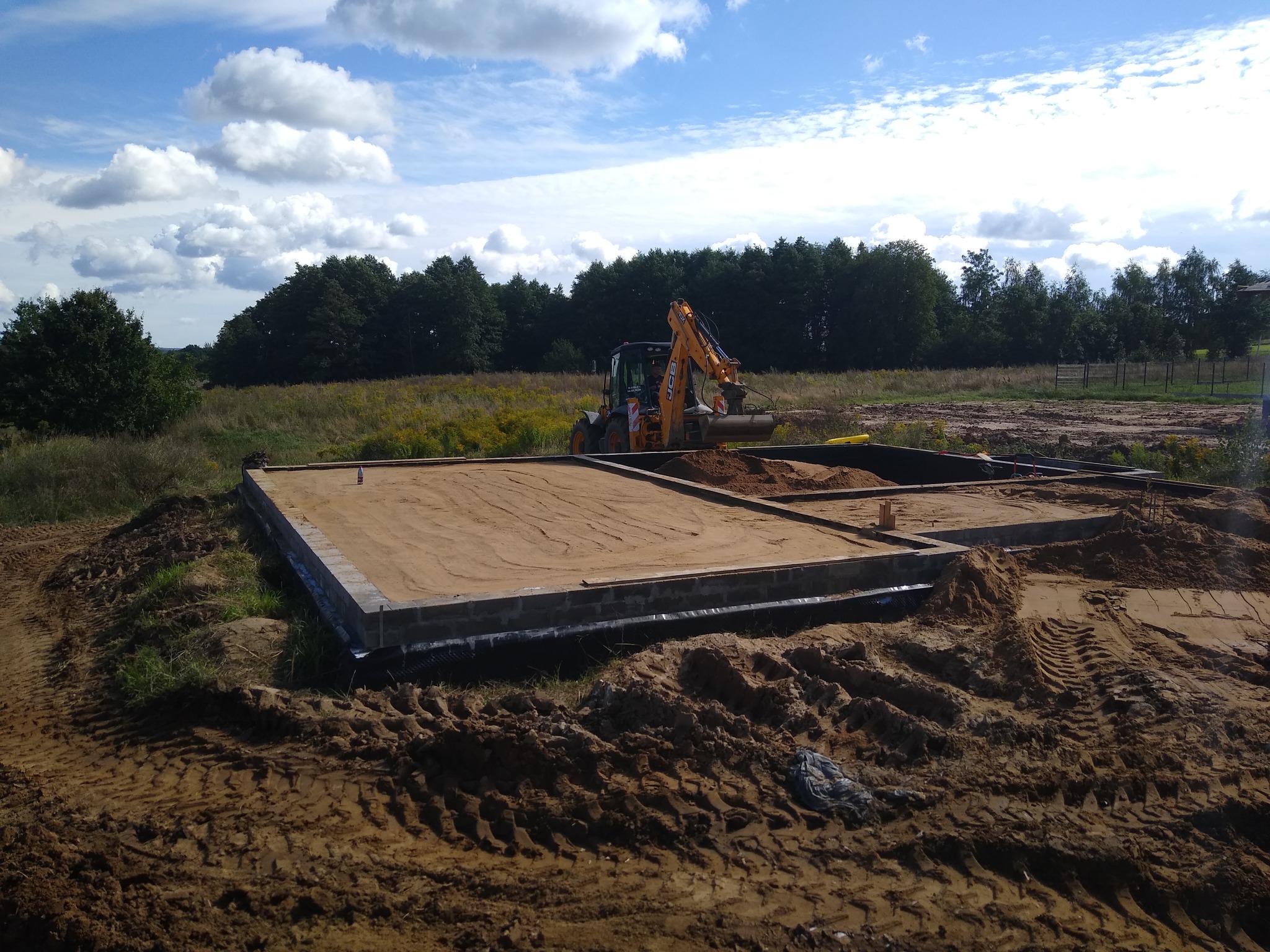 Przygotowanie fundamentów pod budynek jednorodzinny, widoczny zarys fundamentów z bloczków betonowych, ziemia wyrównana, w tle koparko-ładowarka JCB i drzewa.