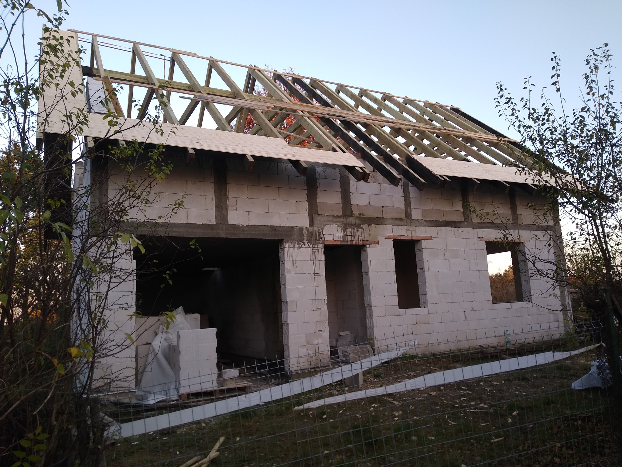 Surowy stan domu z białych bloczków, widoczne otwory okienne i drzwiowe, konstrukcja drewniana dachu w trakcie budowy, otoczenie placu budowy.