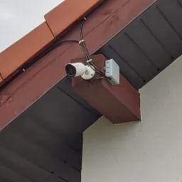 Biała kamera monitoringu zamontowana pod okapem dachu domu jednorodzinnego, przewody przymocowane uchwytami do drewnianej konstrukcji.