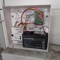 Otwarta skrzynka z elektroniką alarmową, widoczna płyta gł&oacute;wna, zasilacz buforowy marki Europower, okablowanie i narzędzia na dolnej krawędzi.