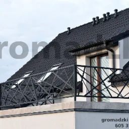 Czarna, geometryczna balustrada balkonowa z metalu na tle dachu z dach&oacute;wki i okna z żaluzjami.