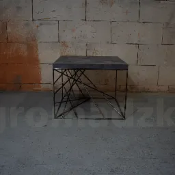 Stolik kawowy z szarego blatu i metalowej podstawy z geometrycznym wzorem, na tle surowej ściany z cegieł