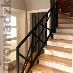 Schody z drewnianymi stopniami i nowoczesną, czarną, metalową balustradą o geometrycznym wzorze, widok z dołu, ściana z cegły w tle.