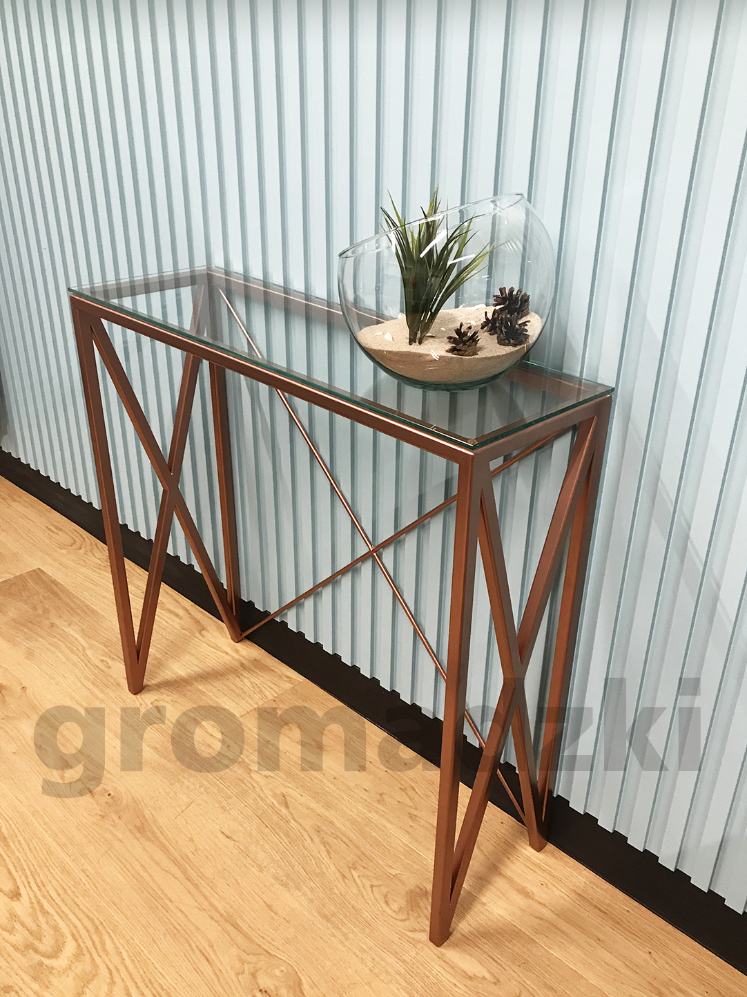 Konsola z metalową podstawą w kolorze miedzi i szklanym blatem, na której stoi szklana misa z piaskiem i roślinami, na tle ściany z pionowymi panelami.