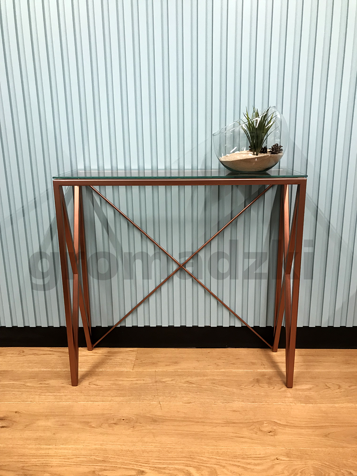 Metalowa konsola z szklanym blatem i dekoracyjną rośliną w szklanym naczyniu, na tle błękitnej, pionowo żłobkowanej ściany.