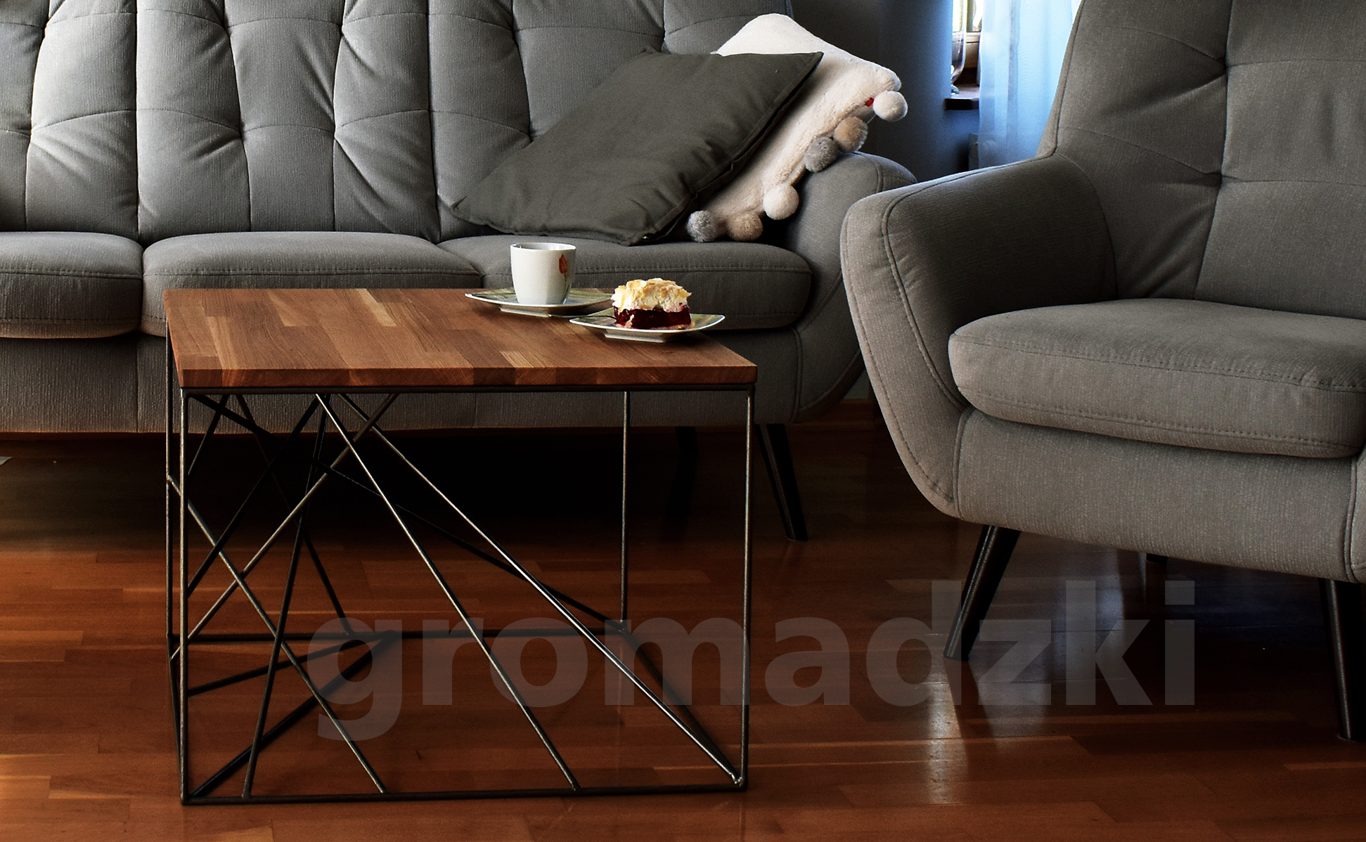 Stylowy stolik kawowy z drewnianym blatem i metalową podstawą w geometryczne wzory, na którym stoi filiżanka i talerzyk z ciastem, w salonie w Gdańsku z szarymi meblami tapicerowanymi.