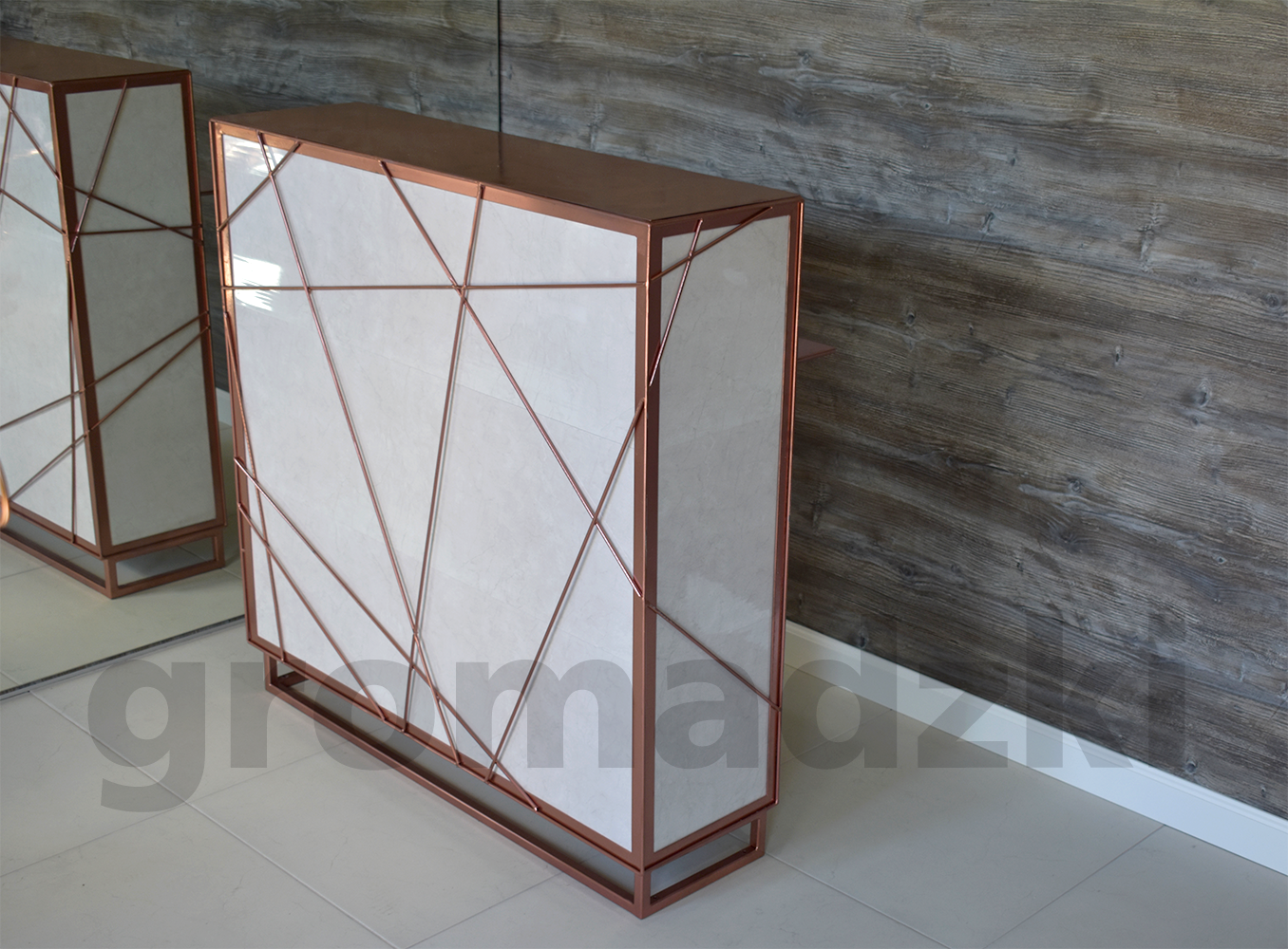 Nowoczesna szafka z białymi frontami i geometryczną ramą w kolorze miedzi, na tle drewnianej ściany i jasnej podłogi.