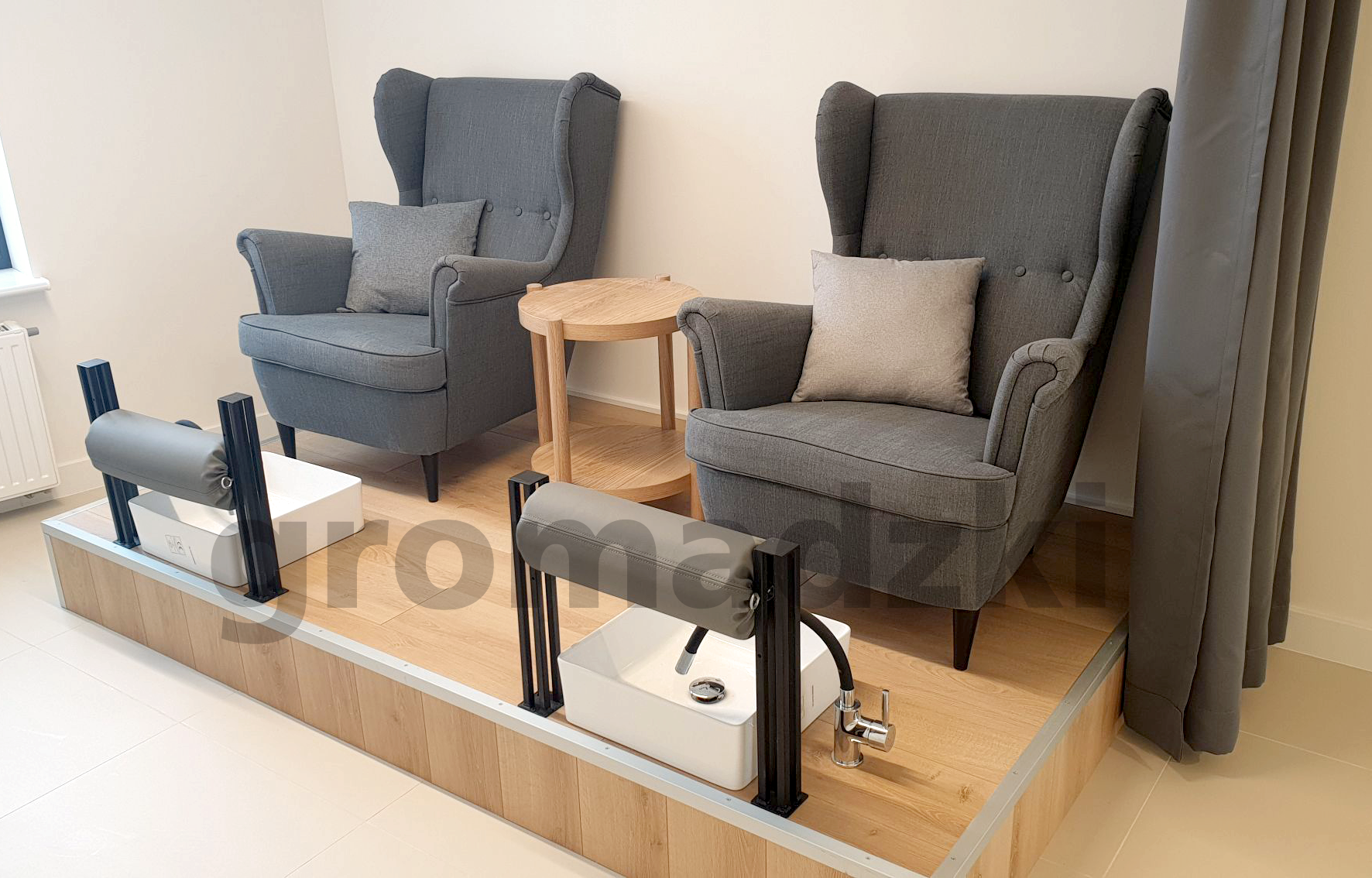 Dwa szare fotele typu uszak z poduszkami, mały drewniany stolik oraz stanowiska do pedicure z białymi misami i chromowanymi kranami, umieszczone na podwyższeniu z drewnopodobnymi panelami.