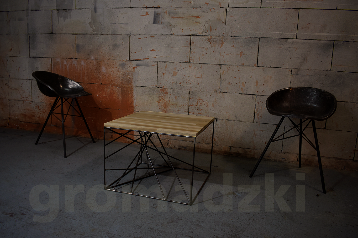 Dwa metalowe stołki barowe i stolik kawowy z drewnianym blatem i metalową geometryczną podstawą na tle surowej ściany z cegieł.