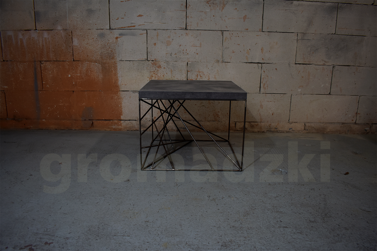 Stolik kawowy z szarego blatu i metalowej podstawy z geometrycznym wzorem, na tle surowej ściany z cegieł