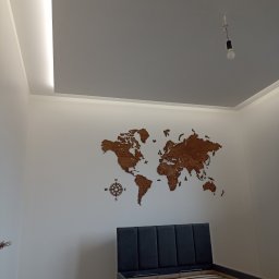 ARTE - prace wykończeniowe - Świeżo wykończona sypialnia z drewnianą mapą świata na ścianie, podświetlanym sufitem i łóżkiem z szarym zagłówkiem, widok na surową żarówkę.
