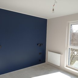 ARTE - prace wykończeniowe - Pomalowane na granatowo ściana z czarnymi włącznikami i kinkietami w surowym pokoju z betonową podłogą, widok na drzewa za oknem.