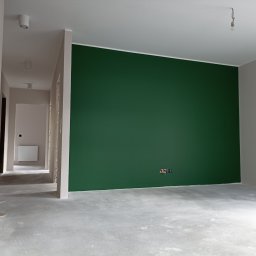 ARTE - prace wykończeniowe - Wnętrze w trakcie malowania: ściana pomalowana na butelkową zieleń, widoczne gniazdka elektryczne, surowa podłoga, w tle korytarz z grzejnikiem.