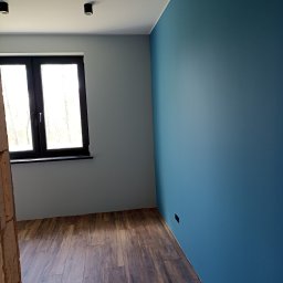 ARTE - prace wykończeniowe - Pomalowane na dwa kolory wnętrze pokoju z drewnopodobną podłogą, widoczne czarne okno i sufitowe lampy.