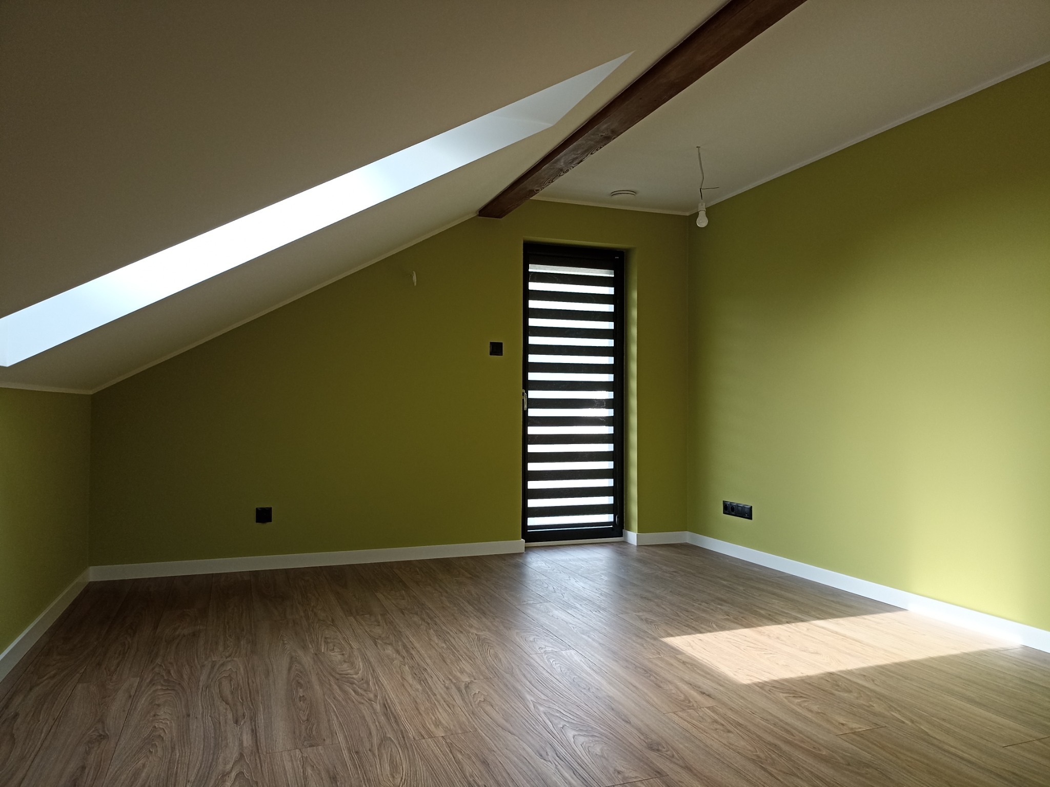 Poddasze po remoncie w Bydgoszczy z oknem dachowym, drewnianą belką stropową i panelami podłogowymi imitującymi drewno, ściany pomalowane na jasnozielony kolor, widoczne gniazdka elektryczne.