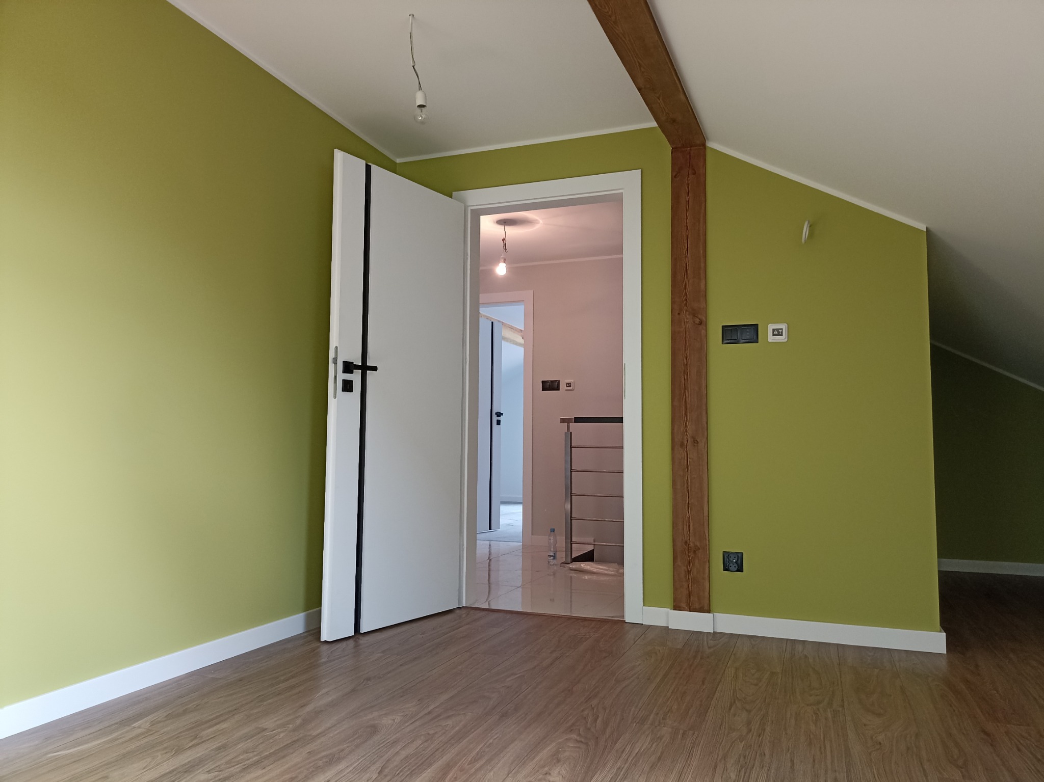 Świeżo pomalowane na oliwkowo poddasze ze skośnymi ścianami, widoczne białe drzwi z czarną klamką, drewniana belka konstrukcyjna i fragment podłogi imitującej drewno.