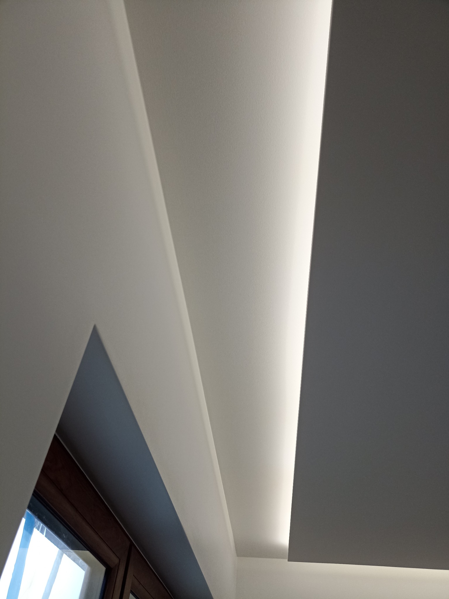 Nowoczesny sufit podwieszany z oświetleniem LED, widok z dołu, fragment okna z drewnianą ramą w lewym dolnym rogu.