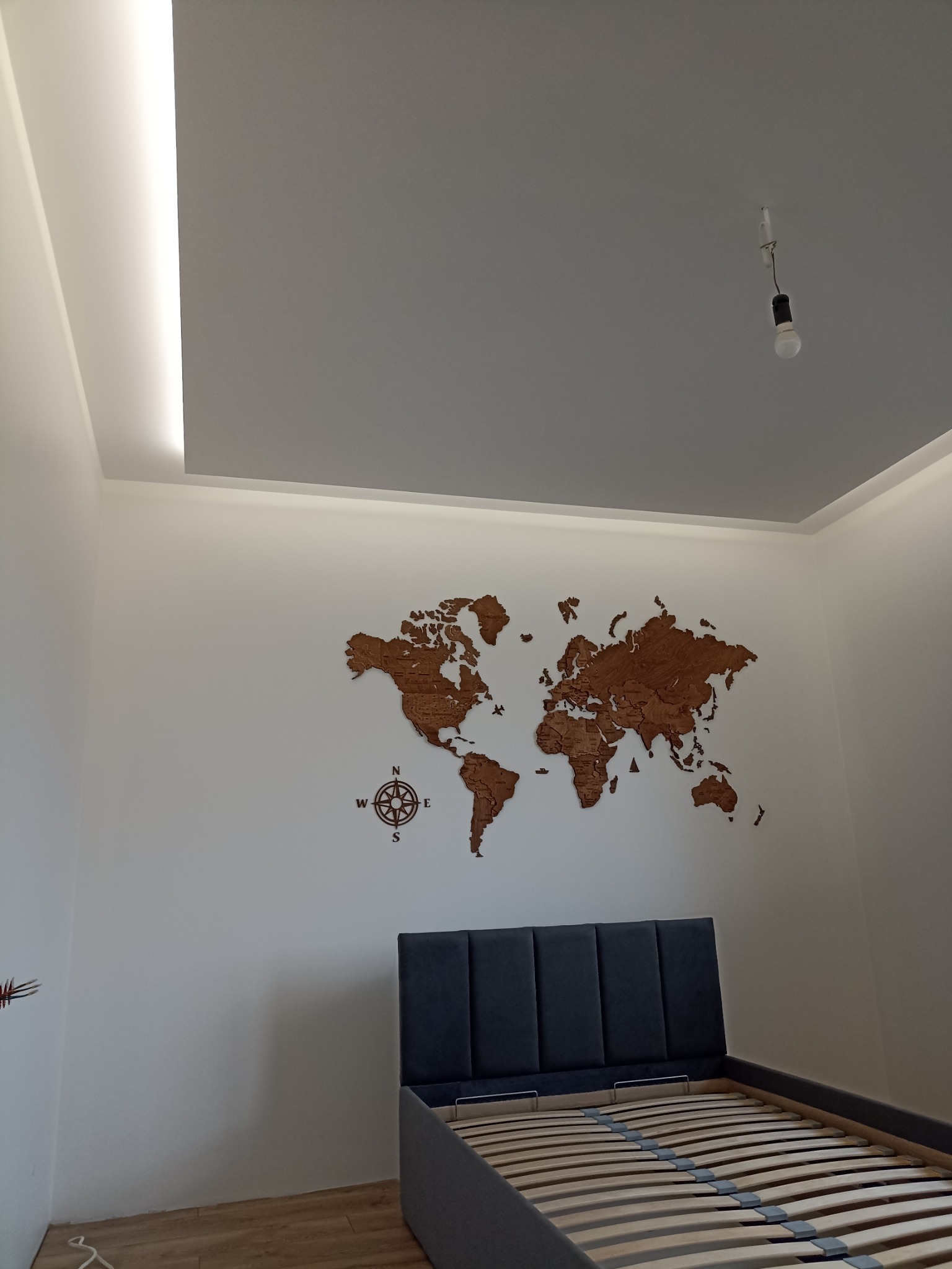 Świeżo wykończona sypialnia z drewnianą mapą świata na ścianie, podświetlanym sufitem i łóżkiem z szarym zagłówkiem, widok na surową żarówkę.