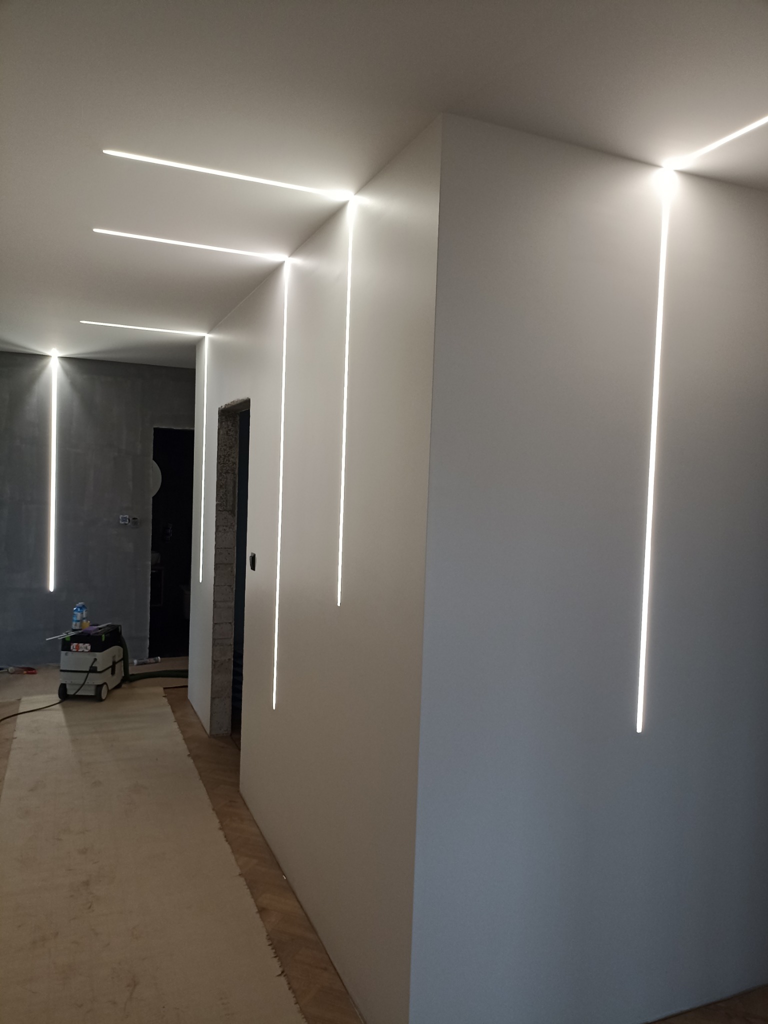 Nowoczesne wykończenie wnętrza korytarza z pionowymi i poziomymi wpuszczanymi liniami LED w ścianach i suficie, widoczny odkurzacz przemysłowy i surowy otwór drzwiowy.
