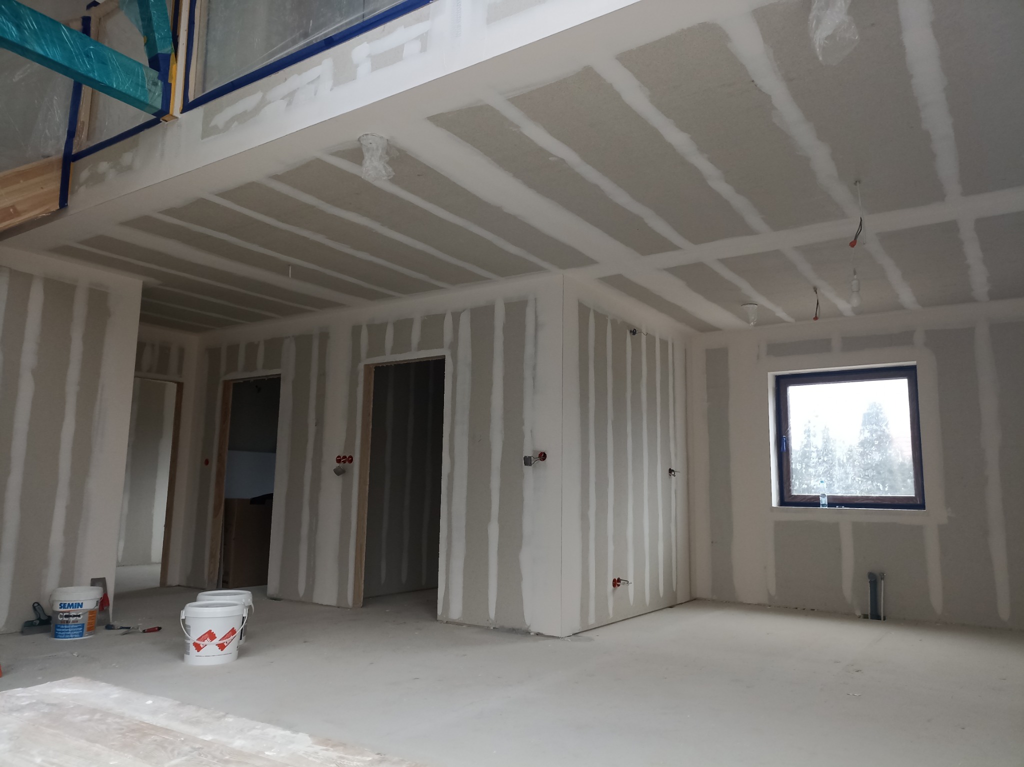 Wnętrze w trakcie prac wykończeniowych: ściany z płyt gipsowo-kartonowych z widocznymi łączeniami, przygotowane do dalszej obróbki, surowe podłoże, puszki elektryczne i wiadro z masą szpachlową.