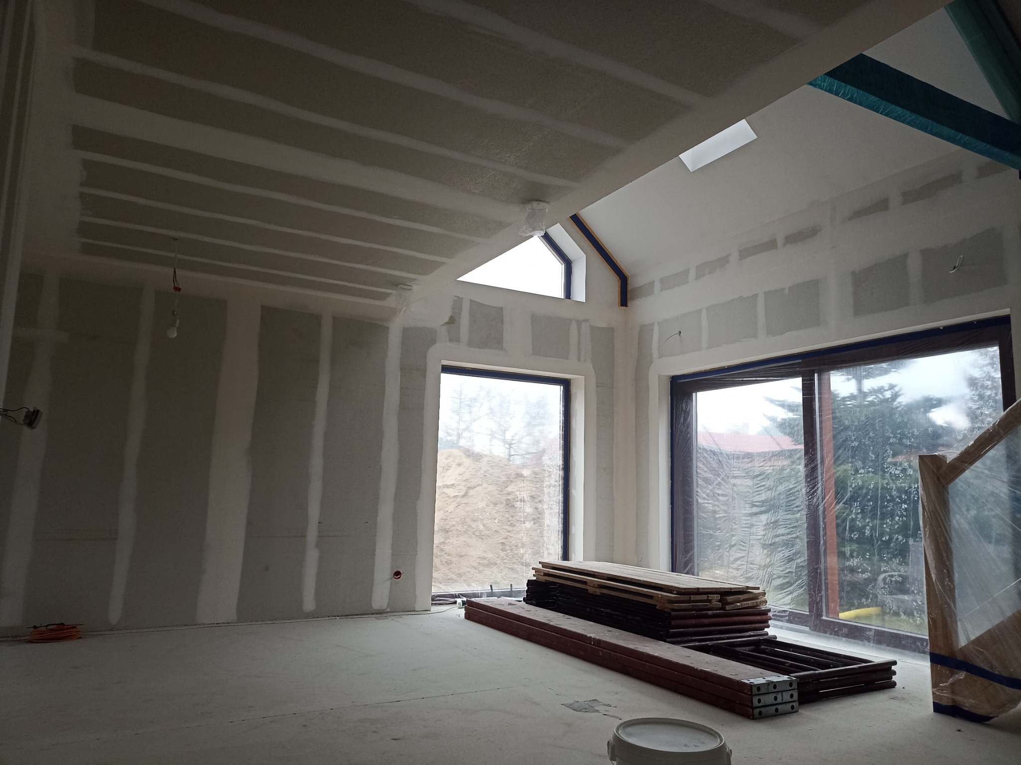 Wnętrze w trakcie prac wykończeniowych: ściany pokryte płytami gipsowo-kartonowymi, widoczne szpachlowanie łączeń, okno dachowe, sterta drewnianych elementów szalunkowych i białe wiadro na podłodze.