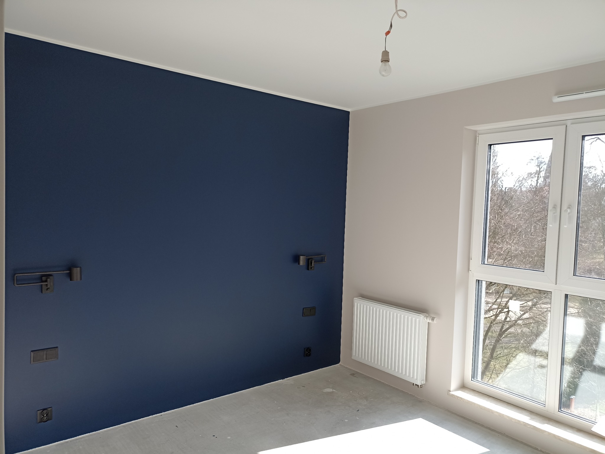 Pomalowane na granatowo ściana z czarnymi włącznikami i kinkietami w surowym pokoju z betonową podłogą, widok na drzewa za oknem.