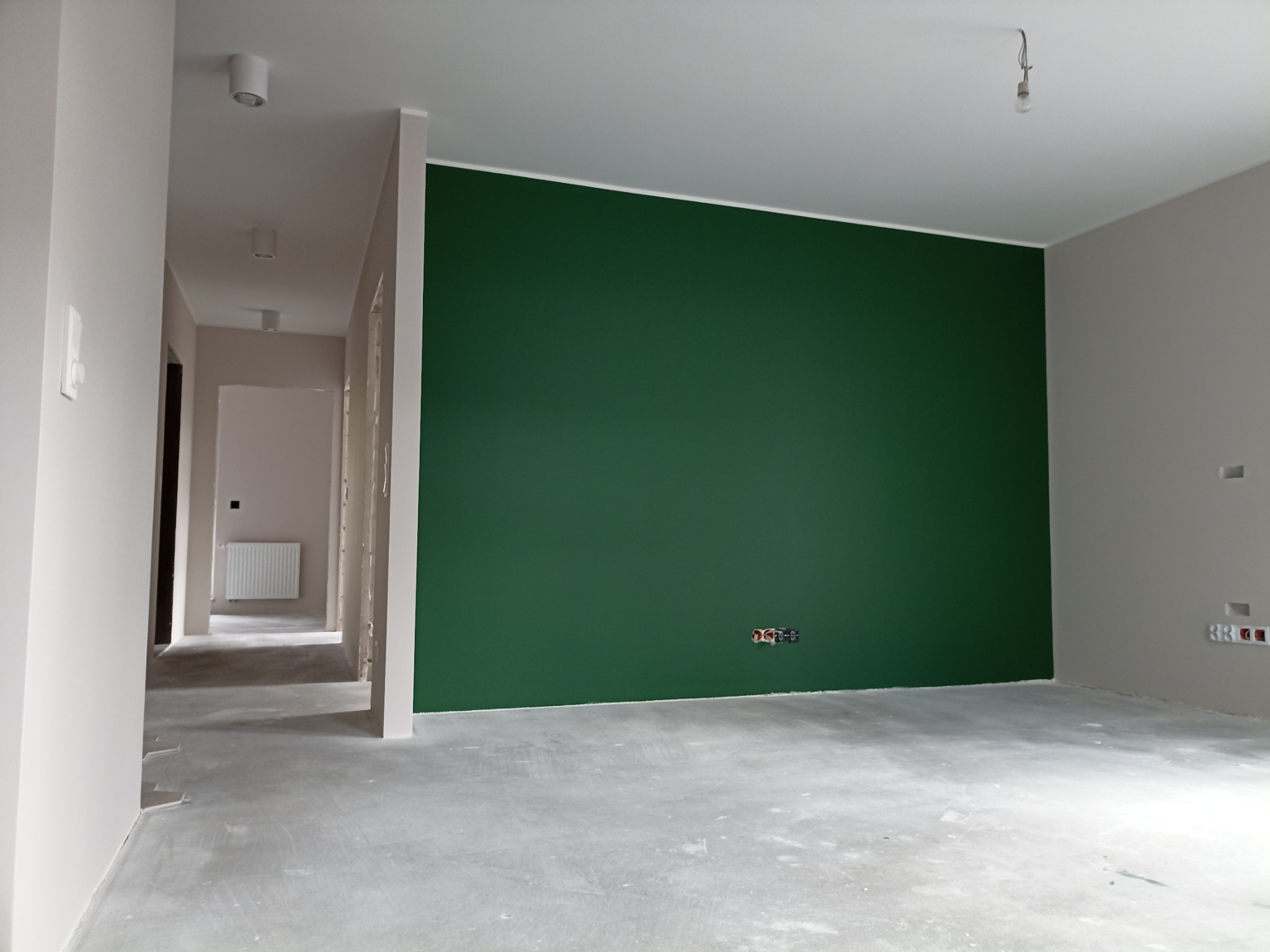 Wnętrze w trakcie malowania: ściana pomalowana na butelkową zieleń, widoczne gniazdka elektryczne, surowa podłoga, w tle korytarz z grzejnikiem.