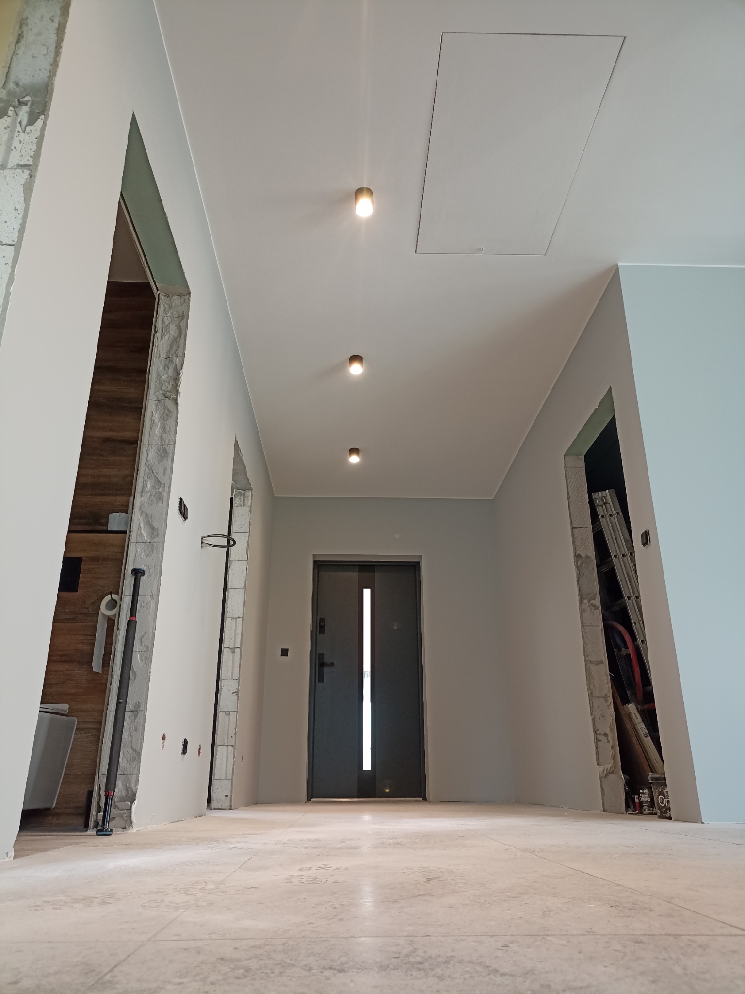 Wnętrze korytarza w trakcie prac wykończeniowych, widoczne surowe ościeża drzwiowe, świeżo pomalowane ściany w odcieniach szarości i beżu, oraz zamontowane oświetlenie punktowe.