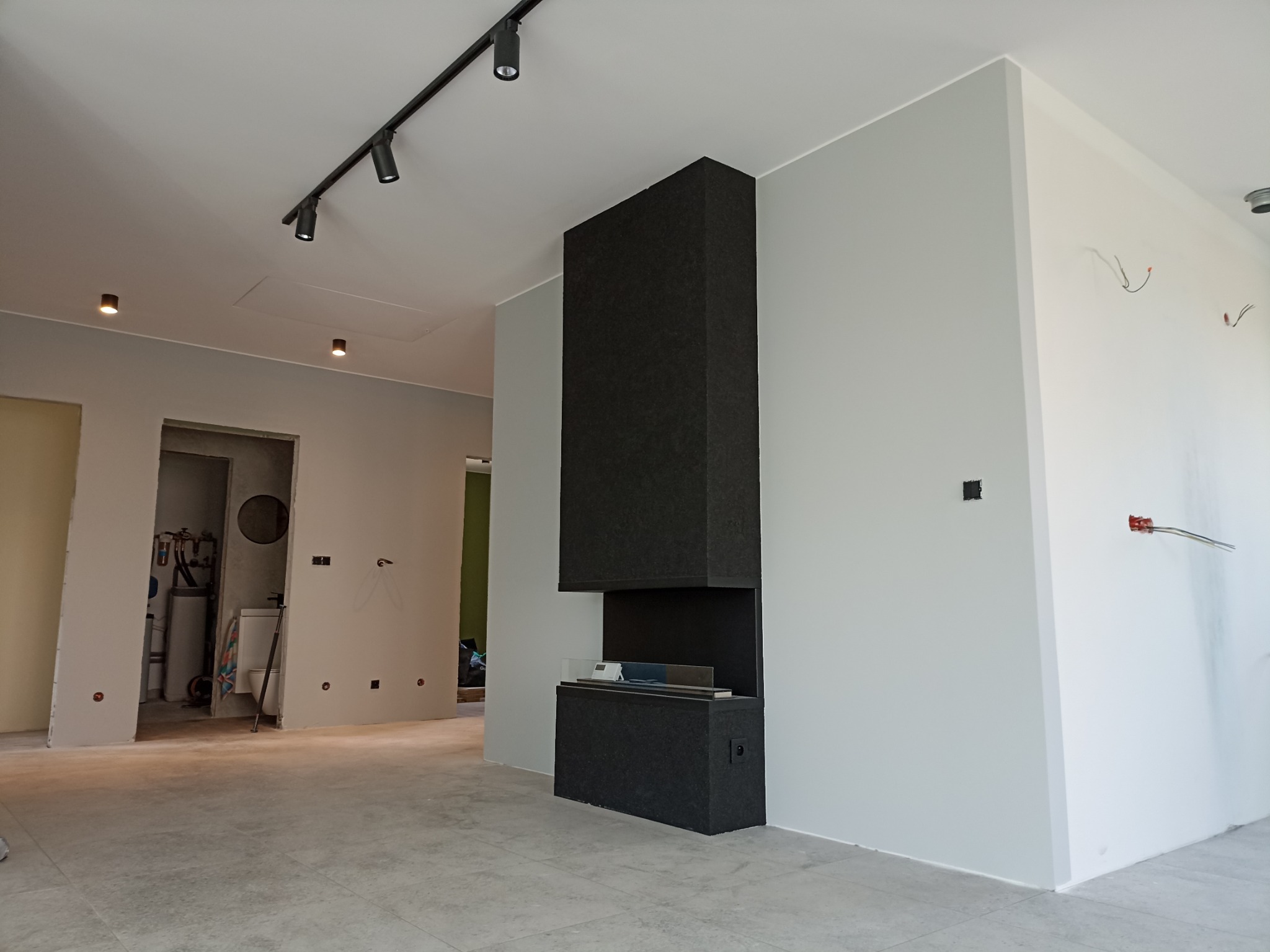 Szare ściany w nowym mieszkaniu z czarnym, nowoczesnym kominkiem bioetanolowym, widoczne niedokończone instalacje elektryczne i surowy stan łazienki w tle.