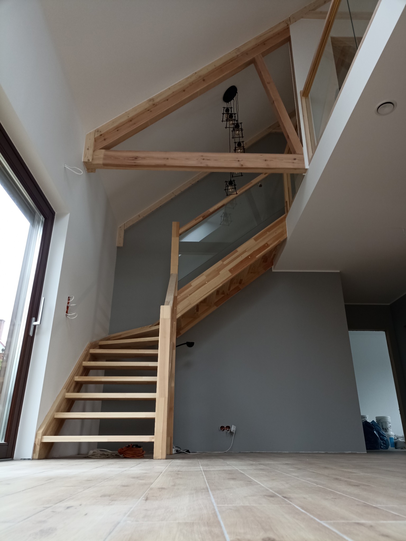 Wnętrze domu z drewnianymi schodami spiralnymi z balustradą ze szkła hartowanego, widoczne drewniane elementy konstrukcyjne dachu i designerska lampa wisząca.