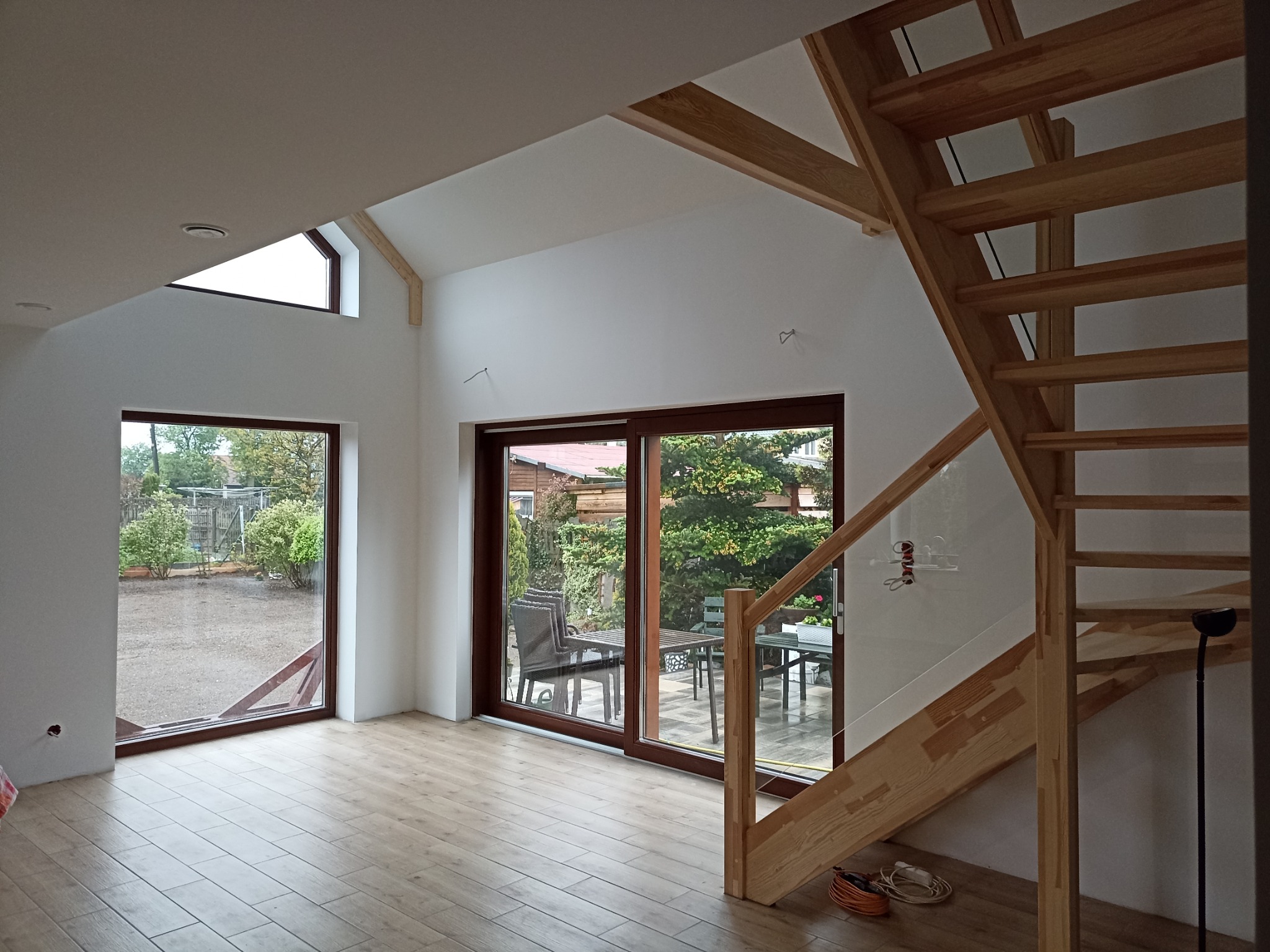 Jasne wnętrze domu z drewnianymi schodami i dużymi oknami wychodzącymi na ogród, widoczne niedokończone prace wykończeniowe.