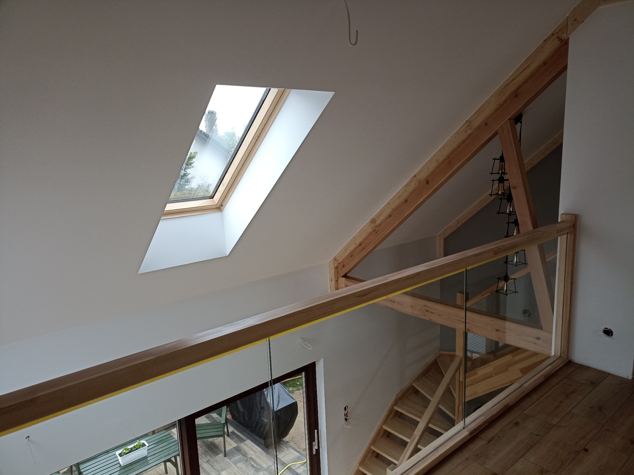 Wnętrze domu w trakcie prac wykończeniowych: widok na drewniane schody i balustradę ze szkła, okno dachowe w białym suficie i odsłonięte drewniane elementy konstrukcyjne.