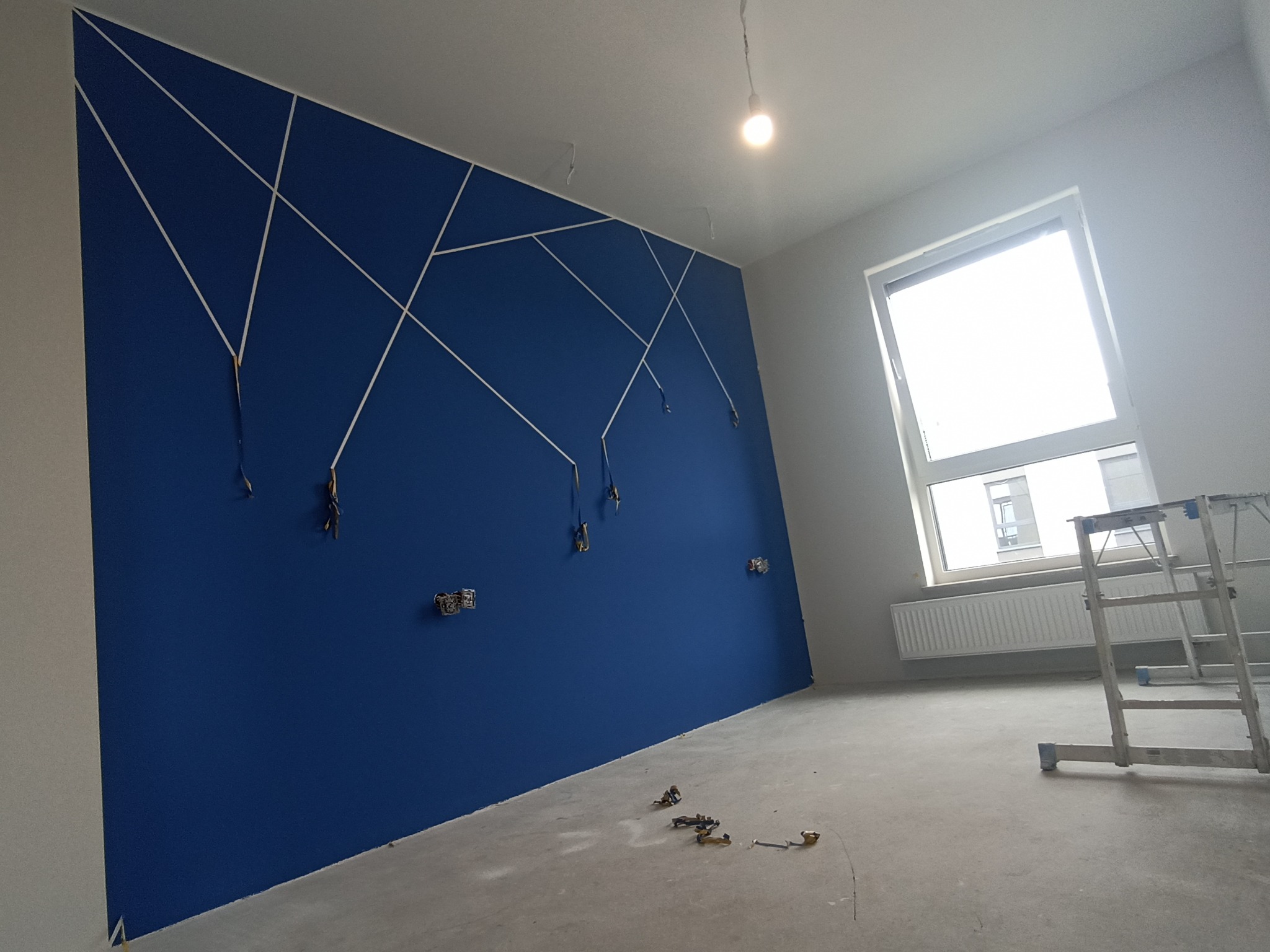 Niebieska ściana z białymi dekoracyjnymi listwami tworzącymi geometryczny wzór, wystające przewody elektryczne, surowy beton na podłodze, drabina w tle, widok okna z budynkiem na zewnątrz.