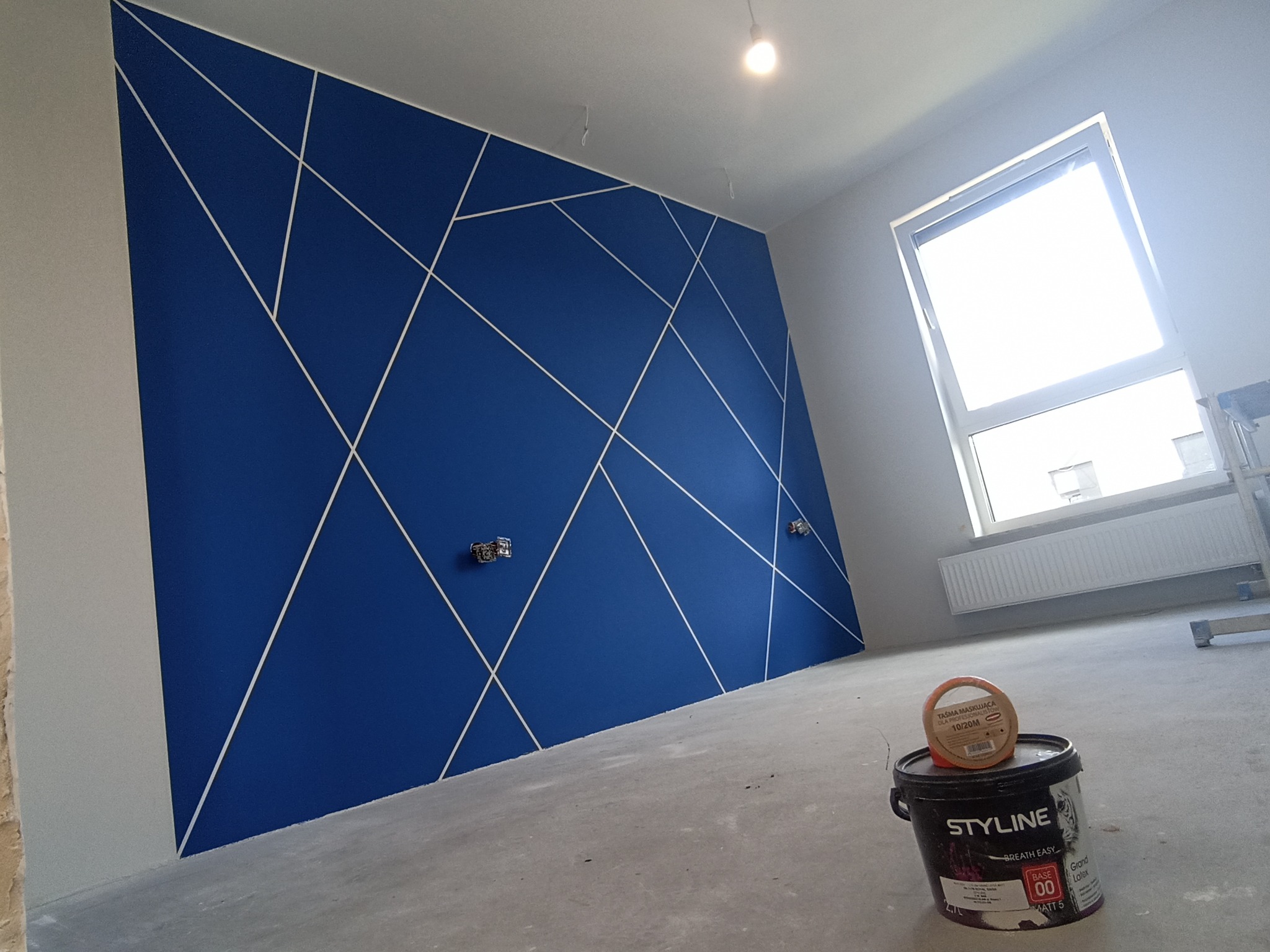 Niebieska ściana z geometrycznym wzorem z białych linii w nowym mieszkaniu, puszka farby 'STYLINE' i taśma malarska na podłodze, okno w tle.