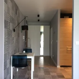 Nowoczesna jadalnia w mieszkaniu: biały stół z krzesłami o przezroczystych oparciach, patchworkowa ściana i podłoga w odcieniach szarości, designerska lampa ścienna z czarnym abażurem.