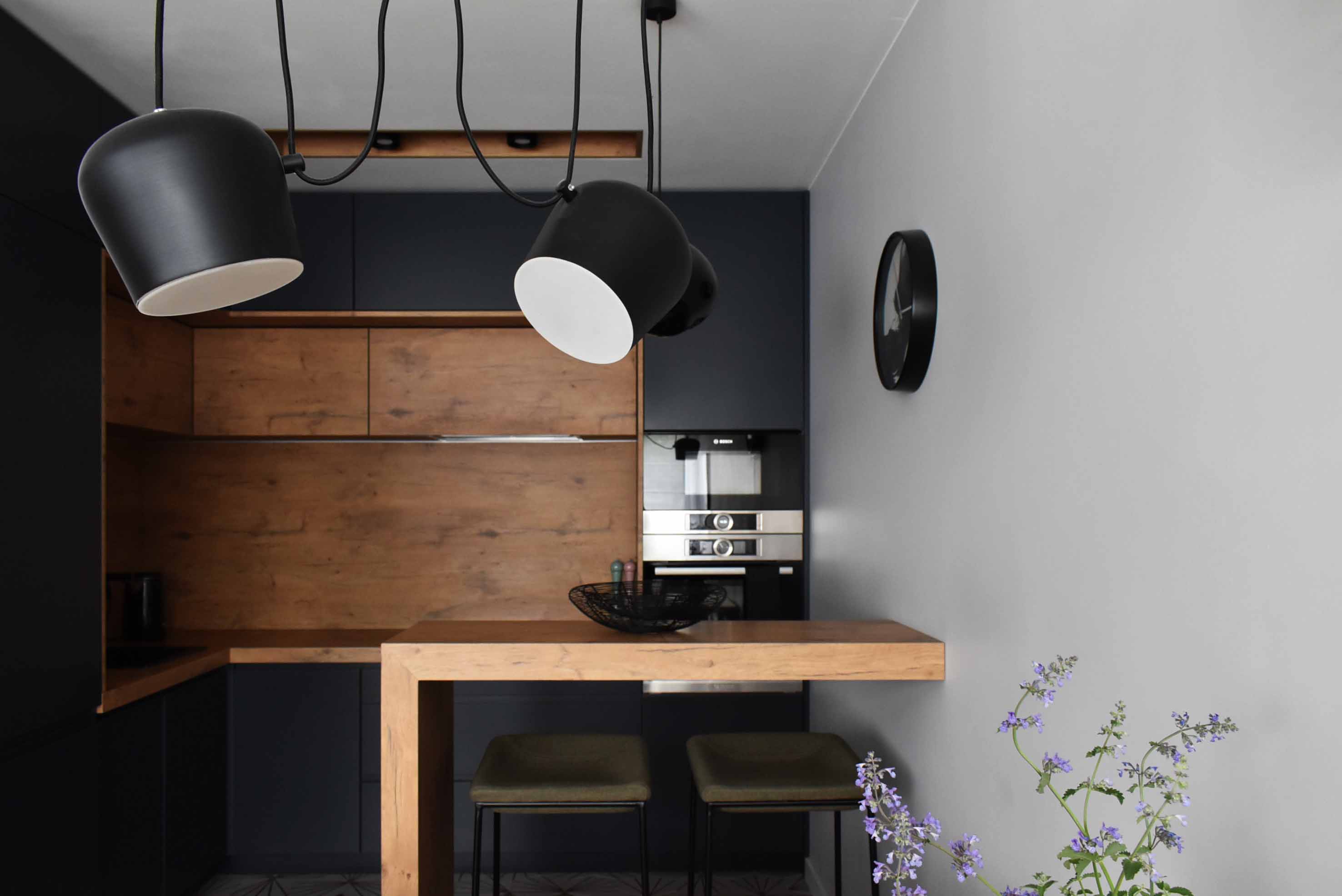 Nowoczesna kuchnia z drewnianymi i granatowymi szafkami, wyspą z drewnianym blatem, czarnymi lampami wiszącymi i kwiatami w wazonie.