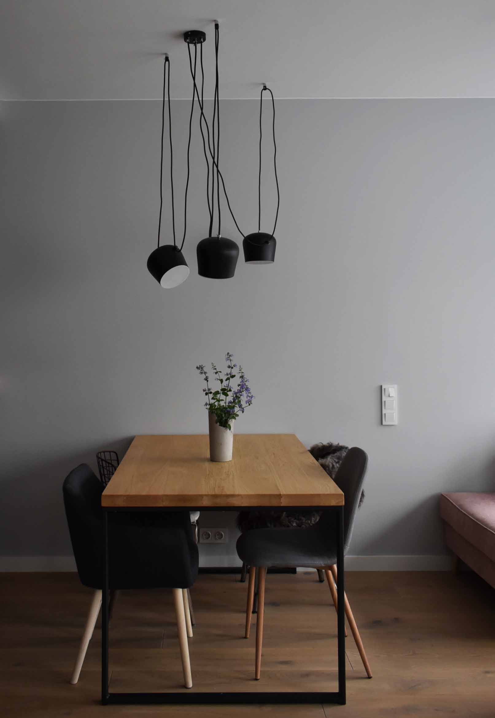 Jadalnia z drewnianym stołem na metalowej podstawie, otoczonym krzesłami z welurową tapicerką w odcieniach szarości i czerni, oświetlona trzema czarnymi lampami wiszącymi z różnymi kształtami...