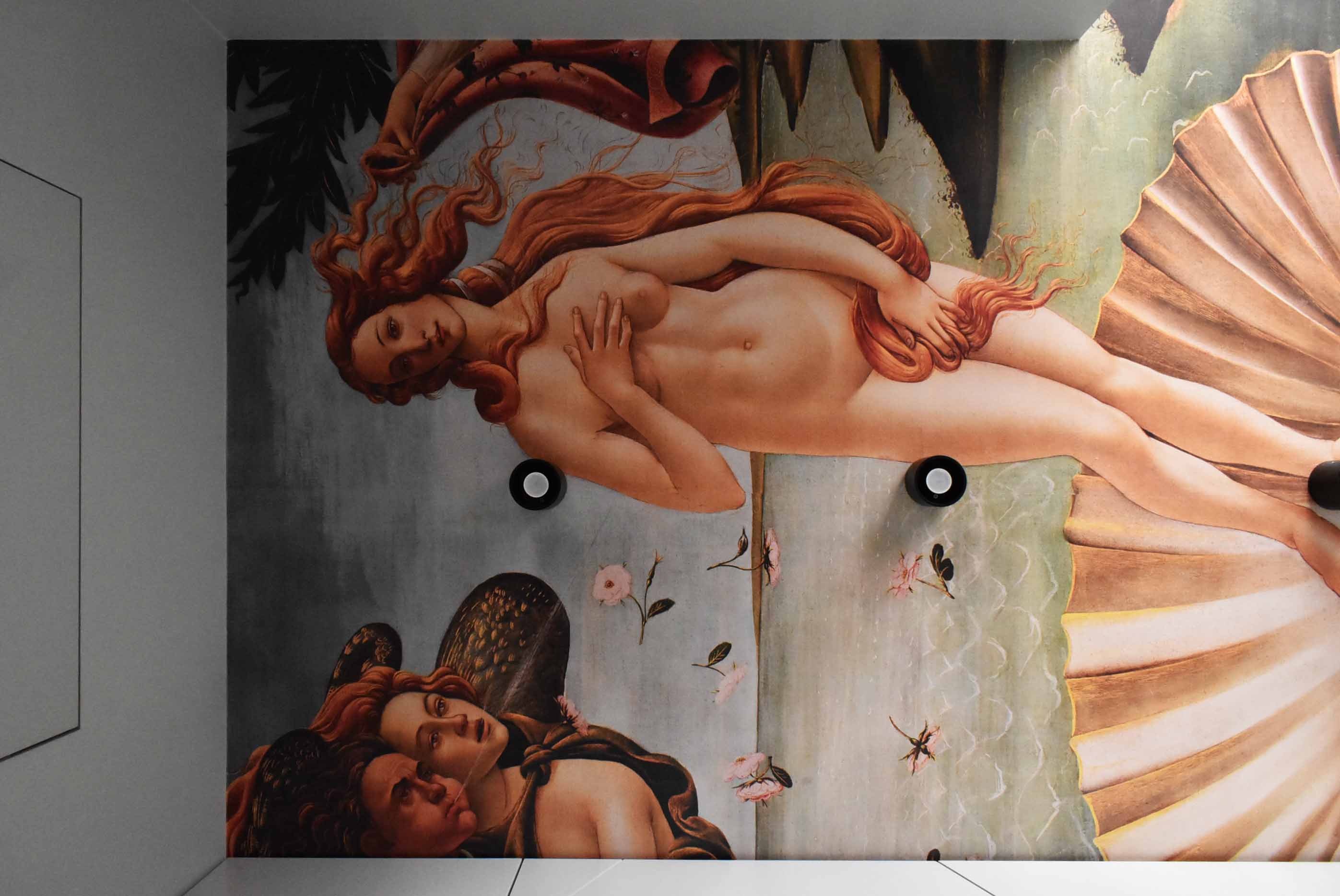 Fragment ściany z reprodukcją obrazu 'Narodziny Wenus' Botticellego, oświetlony dwoma punktowymi lampami sufitowymi.