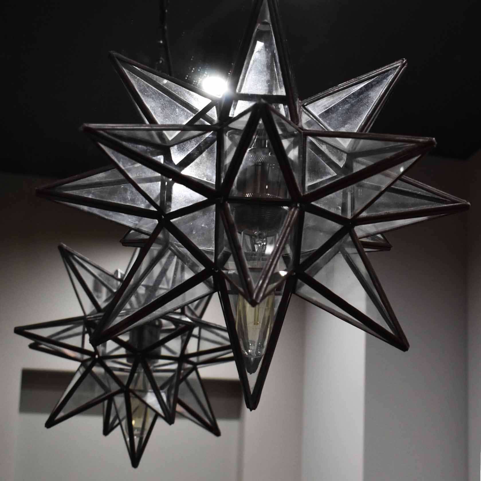 Dwa szklane, geometryczne żyrandole w kształcie gwiazdy, zawieszone na tle ciemnego sufitu i jasnej ściany, oświetlone żarówkami typu Edison.