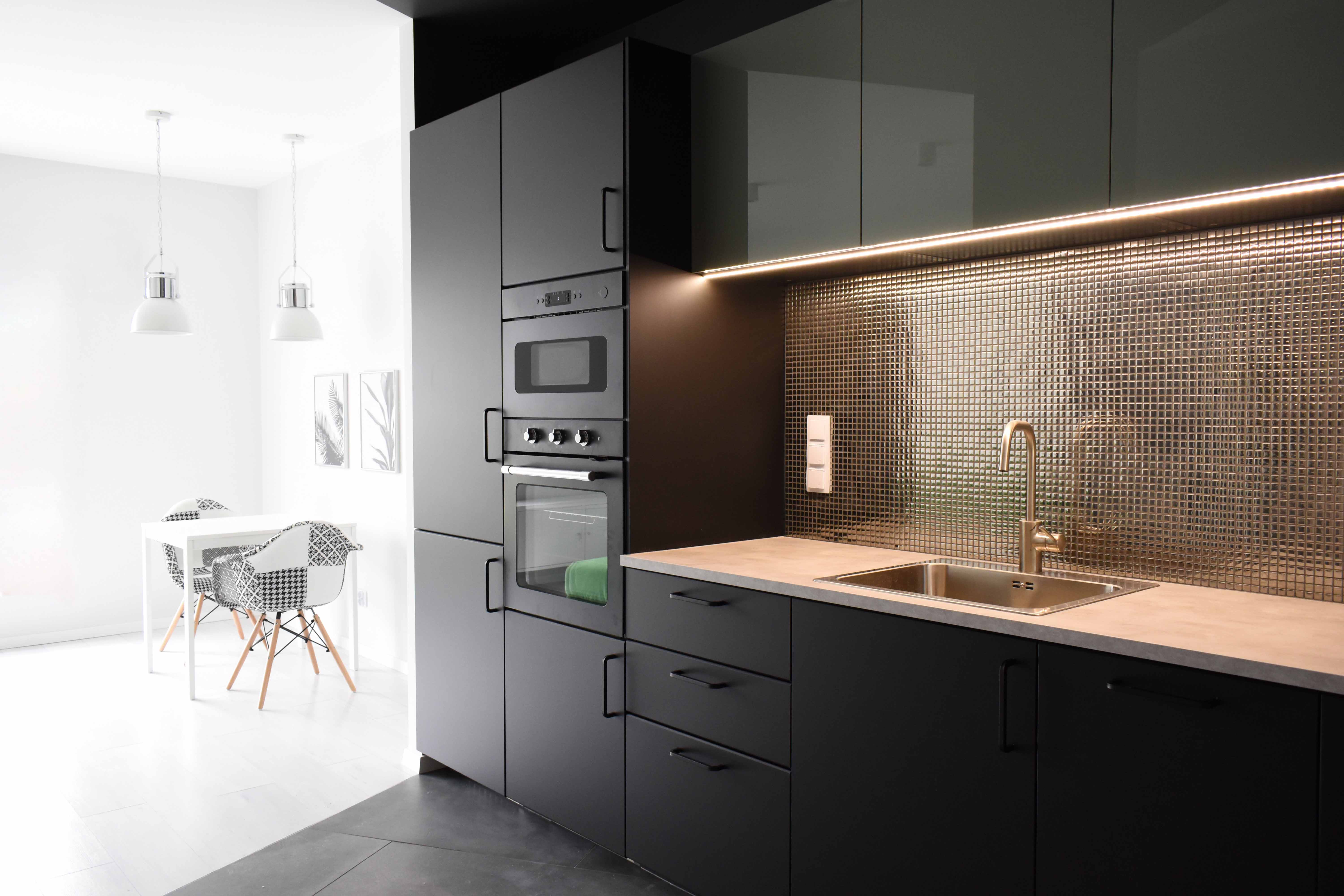 Nowoczesna kuchnia z czarnymi matowymi szafkami, złotą mozaiką na ścianie nad blatem i stalowym zlewozmywakiem z baterią w kolorze mosiądzu, widok na jadalnię z białym stołem i krzesłami...