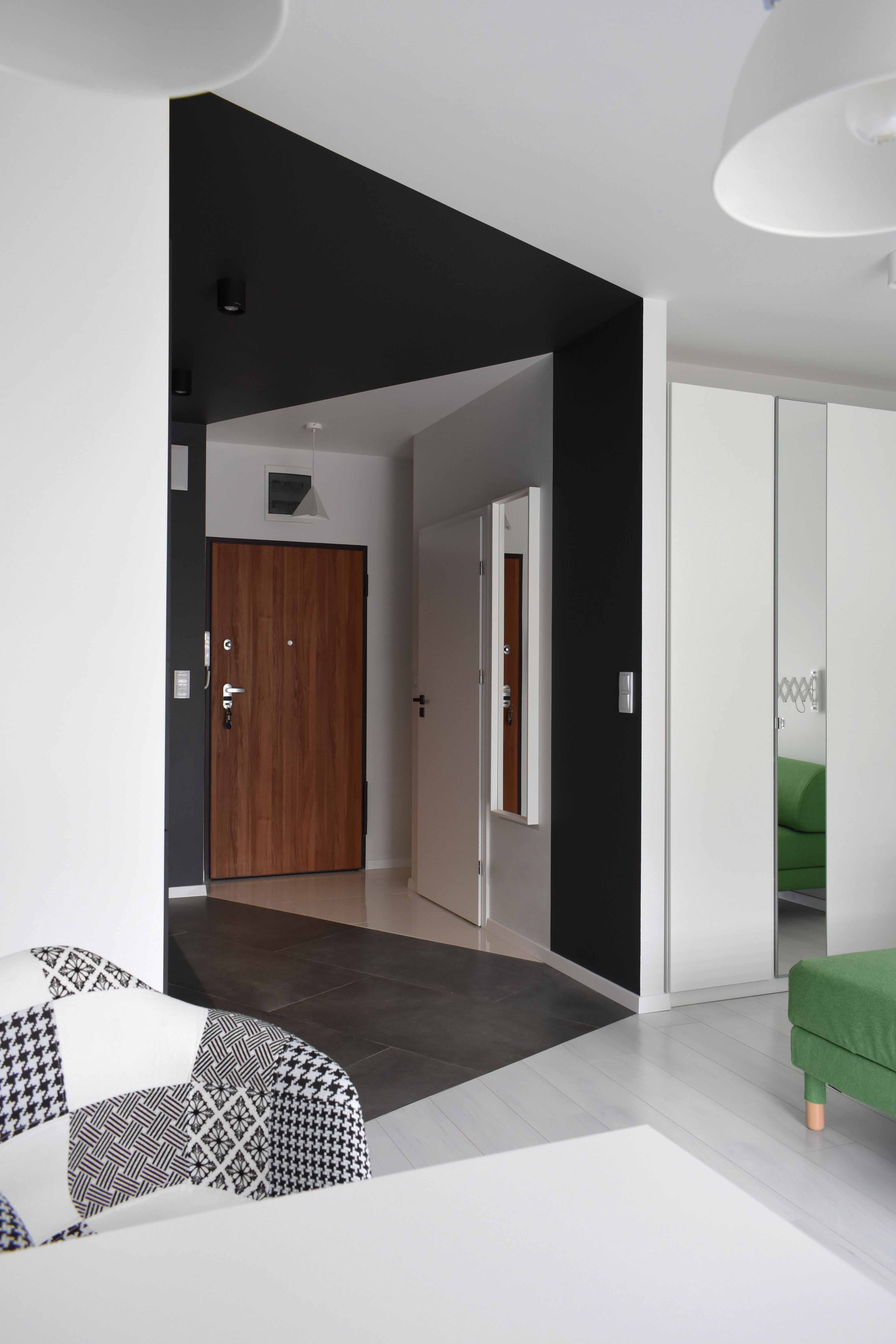 Nowoczesny przedpokój z drewnianymi drzwiami wejściowymi, czarną ścianą akcentującą, szafą z lustrami i patchworkowym fotelem