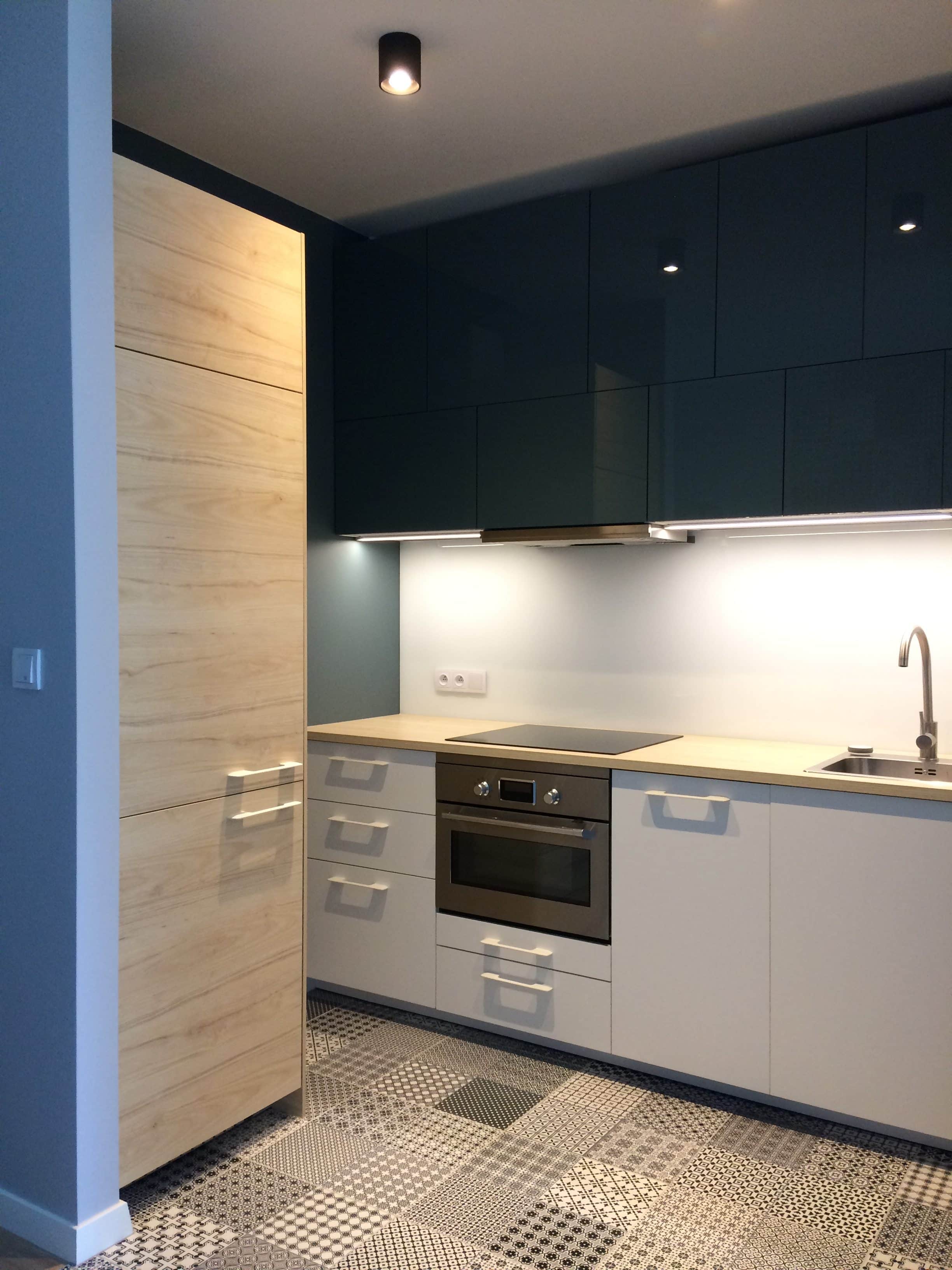 Nowoczesna kuchnia z drewnianymi i granatowymi szafkami, jasnym blatem, srebrnym piekarnikiem i wzorzystą podłogą.