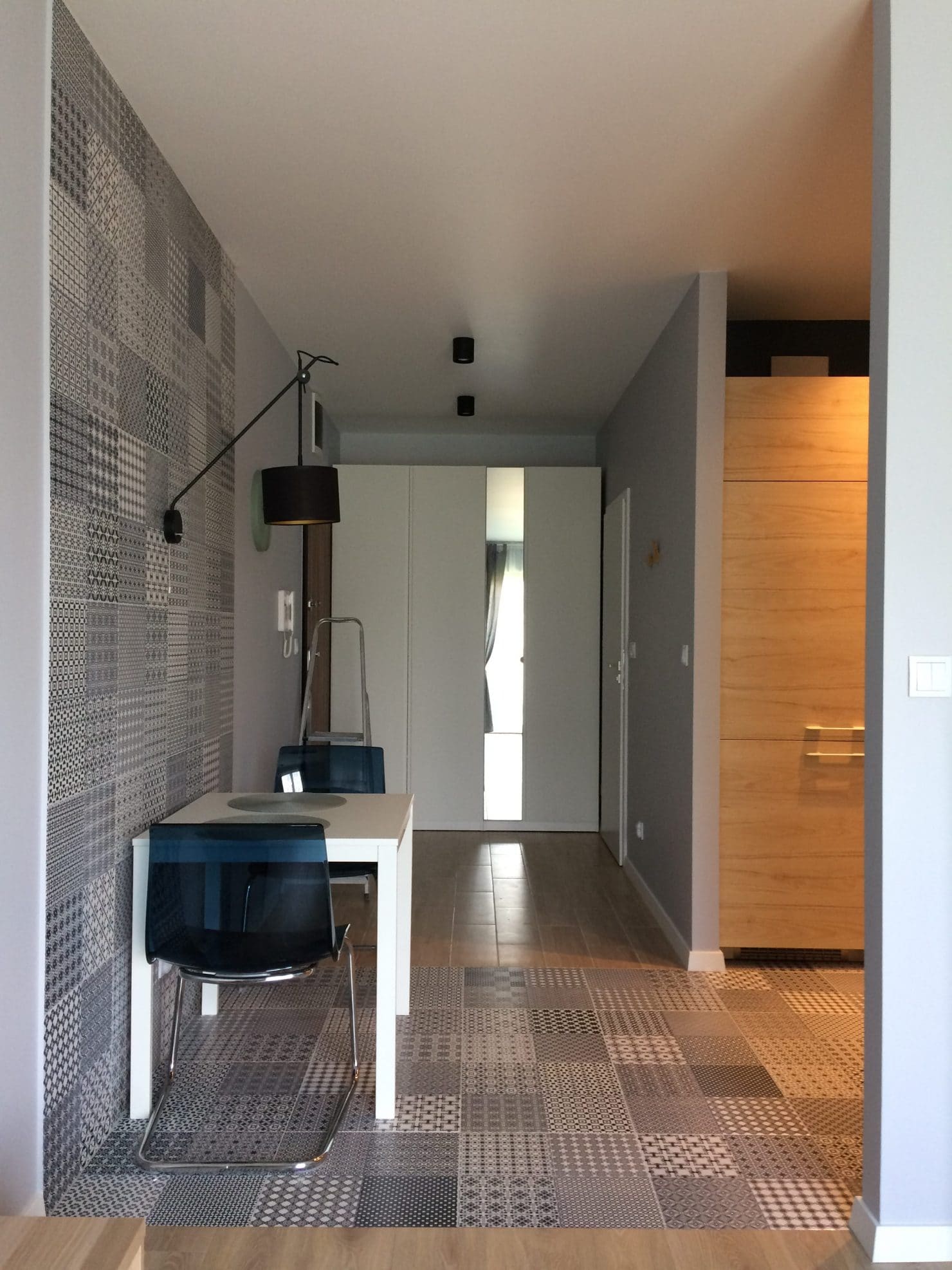 Nowoczesna jadalnia w mieszkaniu: biały stół z krzesłami o przezroczystych oparciach, patchworkowa ściana i podłoga w odcieniach szarości, designerska lampa ścienna z czarnym abażurem.