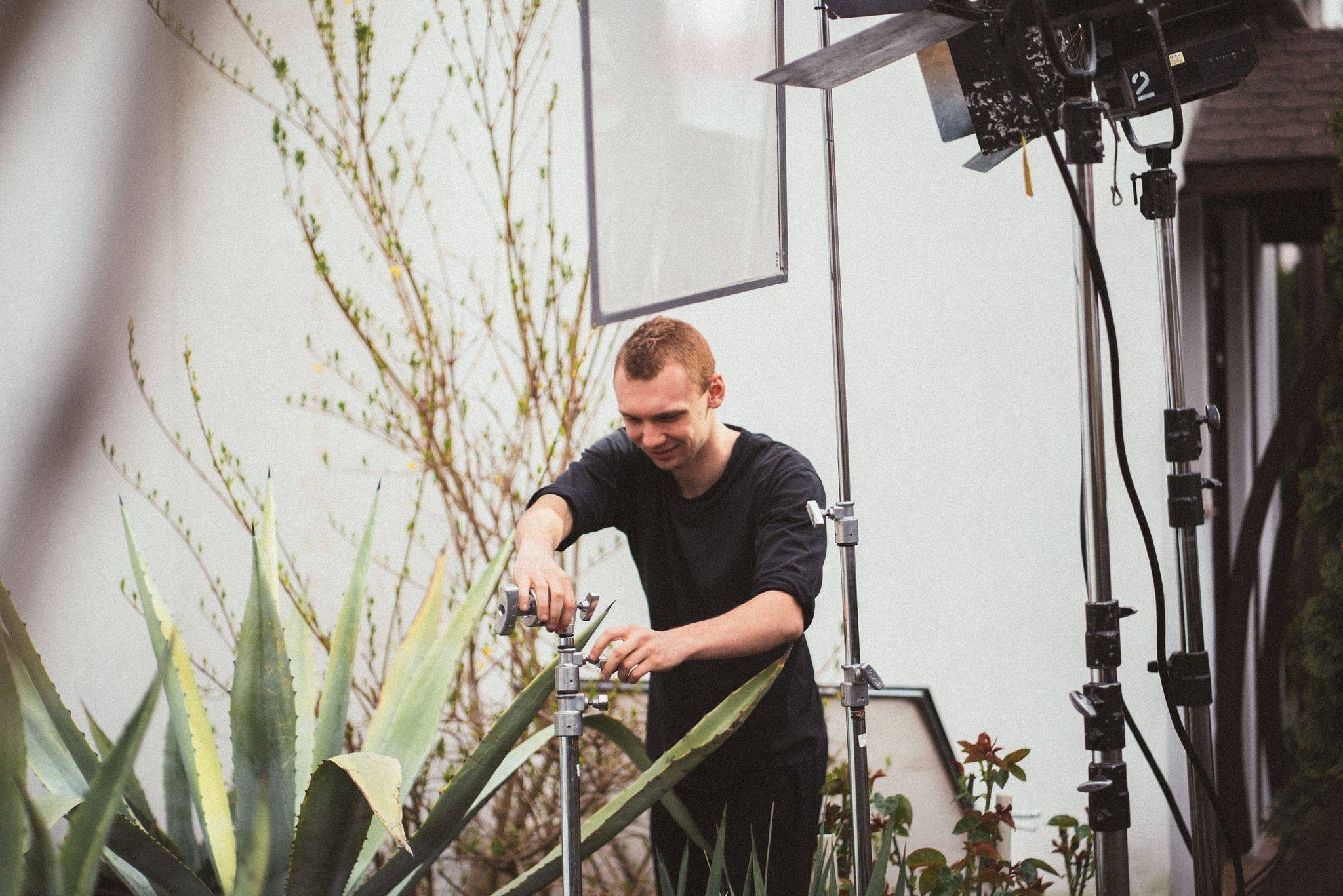 Młody mężczyzna reguluje statyw oświetleniowy w plenerze, z widocznymi roślinami egzotycznymi i krzewami, przygotowując się do filmowania.